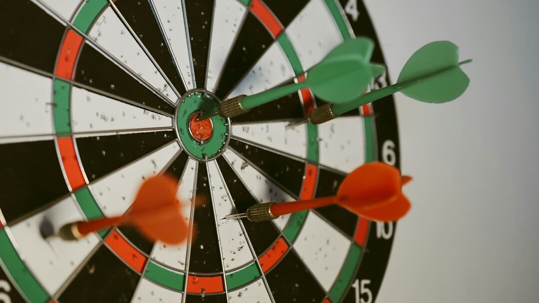 A dartboard with two green darts and one orange dart embedded in it